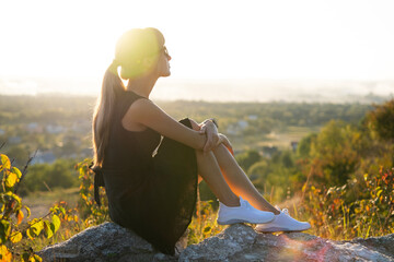 Young pretty woman in black short summer dress sitting on a rock relaxing outdoors at sunset....