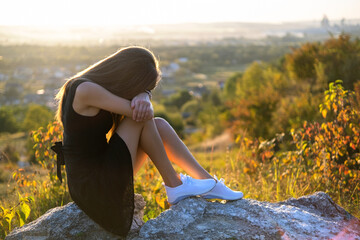 Young depressed woman in black short summer dress sitting on a rock thinking outdoors at sunset....