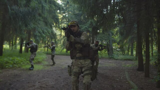 Squad of five fully equipped soldiers in camouflage with assault rifles and machine gun moving forward in forest, weapon in firing position during reconnaissance military mission at daybreak