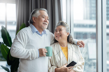 old asian senior couple retired age standing hand hold hot drink look outside window home isolate concept,old senior waiting for lockdown is over home interior background