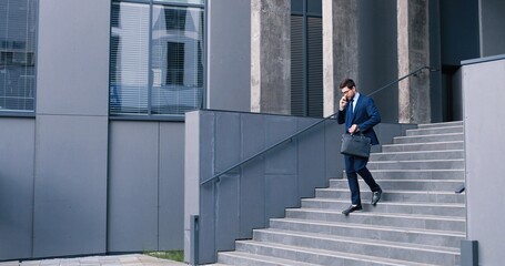 Chatting with pleasure. Full length view of happy young qualified caucasian businessman in suit is going down stairs at the street and holding mobile phone while having conversation with somebody