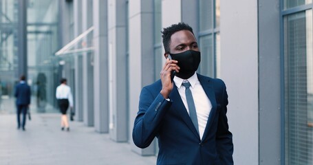 Waist up portrait view of attractive, elegant mature man wearing protective mask walking near the business center. He holding cellular in hand and chatting with pleasure