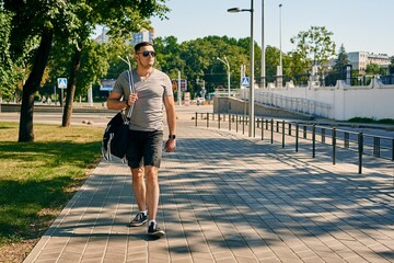 cute athletic brunette guy in sunglasses, shorts and gray t-shirt with a gym bag walks down the street