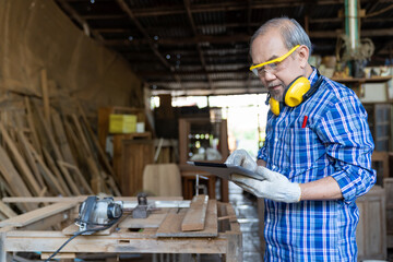 Asian senior male carpenter using digital tablet in carpentry workshop. Carpenter working his job at the workshop