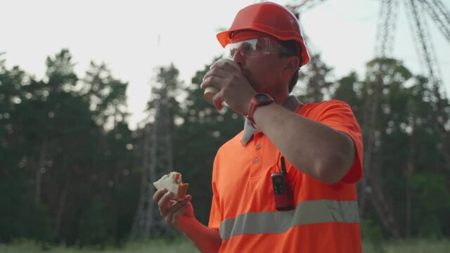 Construction worker on lunch break. Technician engineer man in hard hat eating lunch sandwich and drinking coffee with power line infrastructure at workplace. Electrician eats snack and drinks coffee.