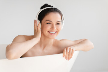 Young happy relaxed woman resting in hot bath shower listening to the music in headphones at home....