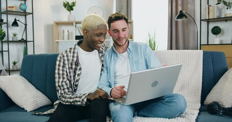 LGBT-community concept where good-looking happy smiling mixed race gay couple in love spending their leisure at home and use computer