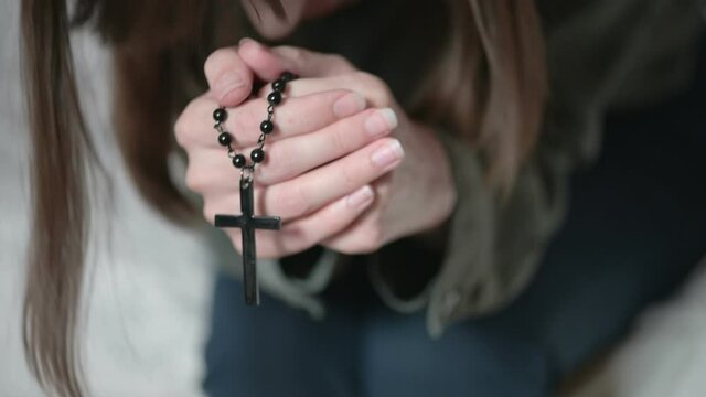 Depressed lady brunette sits on grey sofa and prays holding black rosary in hands and kissing cross at home in morning closeup.