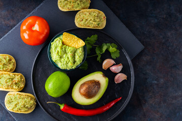 Guacamole sauce, nachos chips and ingredients on a dark background. Mexican guacamole sauce with avocado, ingredients and corn nachos. Copy space. Top view