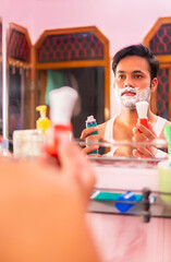 Indian young man looking in to mirror and applying shaving cream on his facial hair and shaving at home.