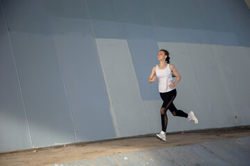Fototapeta na wymiar A young woman in a white T-shirt and black pants runs under the bridge in the evening. Running trainings in the city. Preparing for the competition.