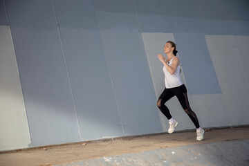 Fototapeta na wymiar A young woman in a white T-shirt and black pants runs under the bridge in the evening. Running trainings in the city. Preparing for the competition.
