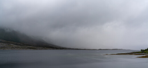 panorama landscape view of a fjord with mystical fog and cloud cover coming down from the mountains over the ocean