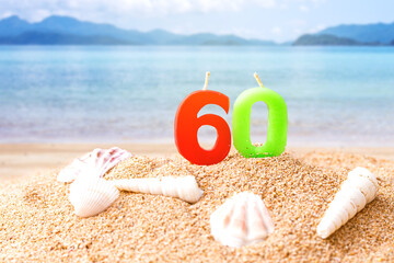 candle number 60 and sea shell over blurred blue sea and sand beach with clear blue sky on day noon light.