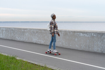 Active urban lifestyle: happy african american girl in warm clothes riding longboard at seaside road. Smiling young woman on skateboard. Skateboarder first ride after winter season. Freedom concept