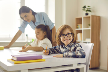 Portrait of funny little child in glasses photographed while writing on paper sheet at school desk. Happy elementary student sitting in classroom, holding pen or pencil, smiling and looking at camera