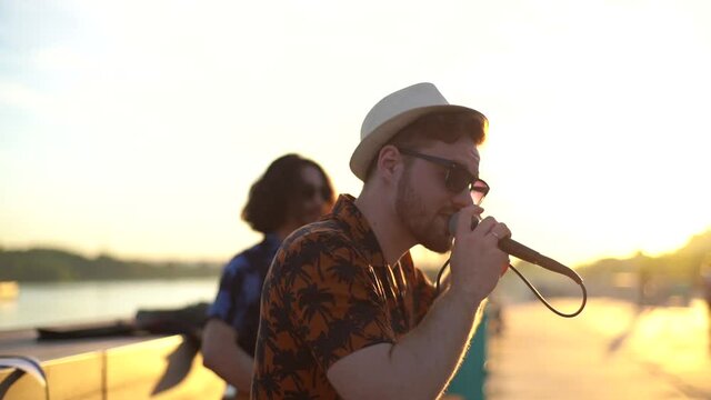Close-up side view of young musical band of male creative singer and male guitarist give performance for public in city street, during sunset with bright sunlight.