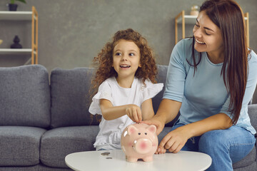 Little child learning to save up and value money. Portrait of happy joyful young mother and her cute daughter sitting on sofa at home, smiling and putting coins in piggy bank. Family finance concept