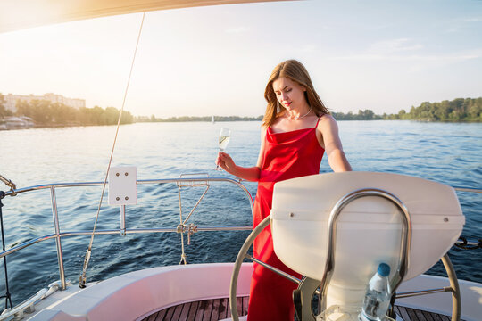 The elegant attractive woman in a long red dress holding a glass of champagne standing on a yacht and looking for river sunset view