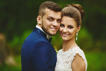 A happy couple of newlyweds hugging and looking in the camera. Beautiful model girl in a wedding dress and handsome man in trendy blue suit