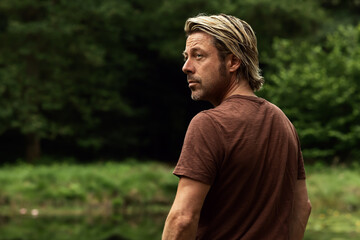 Blonde man in brown t-shirt stands in a forest near a pond. Side