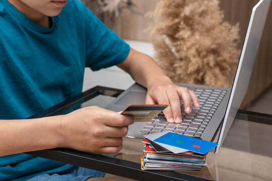 Close up image of boy's hands with credit card and big stack of different cards near laptop executing online payment. New reality, convenience on online shopping from home