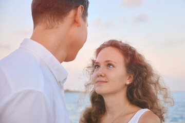 Young adult curly haired woman looks to her beloved man opposite sea. Young adult heterosexual couple at outdoor date in the evening. Attractive young woman looks at a man with a loving look