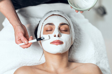 Cosmetologist applying the alginates facial mask to woman while lying on a stretcher in the spa...