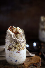 Healthy overnight oatmeal, Bircher muesli, served with bananas, brown sugar, pecan nuts, and maple syrup over a rustic table background. Selective focus with blurred background.