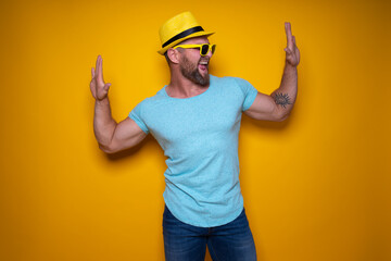 Happy male in blue shirt, sunglasses and yellow hat celebrating success standing on yellow background. 