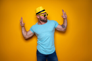 Happy male in blue shirt, sunglasses and yellow hat celebrating success standing on yellow background. 