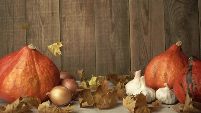 Pumpkins and onion with garlic in autumn composition among falling dry foliage in slow motion. Still-life made of raw food on wooden background. Rustic composition made of vegetables and maple leaves.