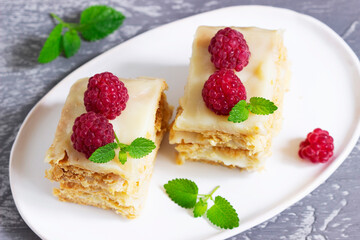 Pieces of Napoleon cake decorated with raspberries and lemon balm leaves on a light background.