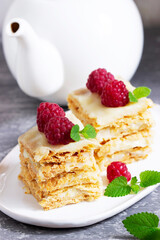 Pieces of Napoleon cake decorated with raspberries and lemon balm leaves on a light background.