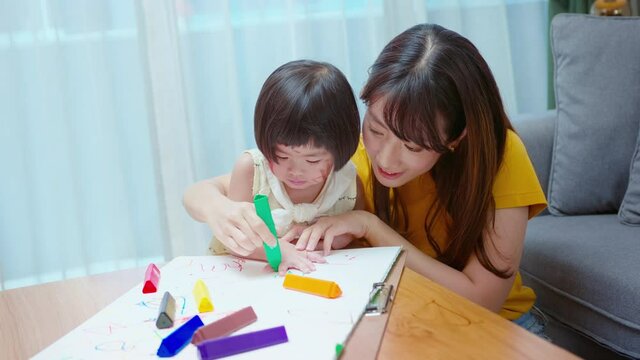 Young mom helping daughter drawing with colored pencils in living room at home.