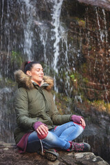 Young woman meditating next to a waterfall in a forest, relaxing in warm clothes, enjoying autumn time isolated in nature, all by herself