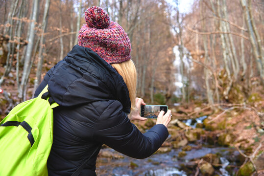 Young woman, a hiker or a backpacker, walking through the forest, taking images and photographs of winter nature, cold river and autumn leaves