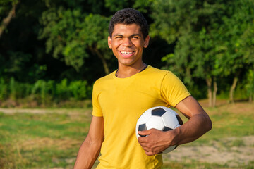 Young smiling Brazilian man looking at the camera holds a ball before playing on a grassy sandy beach in summer. 