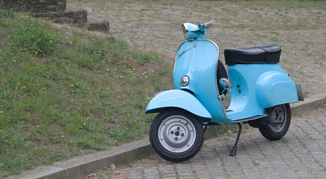 Mota azul claro parada numa rua de uma aldeia antiga, lambreta, fabrico italiano, modelo cl&aacute;ssico dos anos 60, ao lado jardim com flores