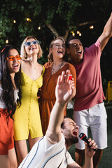 Interracial friends with plastic cup singing near man with microphone during party outdoors