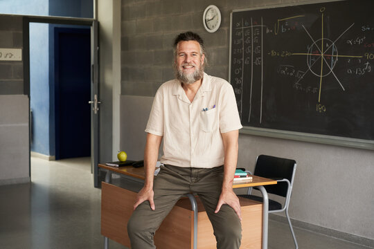Middle-aged bearded male teacher looking at camera in the classroom. Happy and engaged teacher.