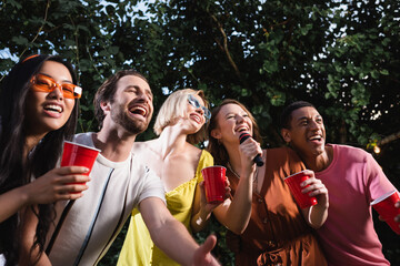 Cheerful multiethnic people singing karaoke and holding drink in plastic cups outdoors