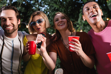 Cheerful multiethnic friends with plastic cups singing karaoke outdoors