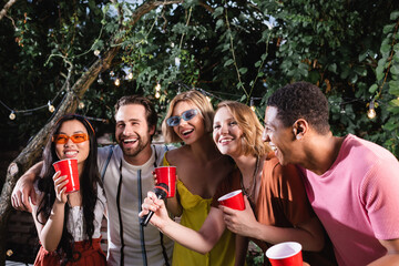happy multiethnic friends with plastic cups singing karaoke in night garden