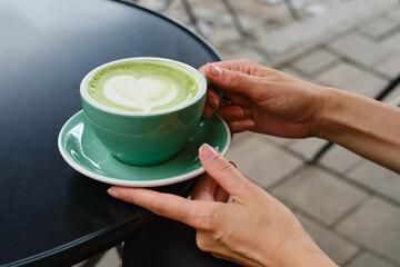 Woman with a cup of matcha latte sits on the summer terrace of a cafe. Delicious green matcha tea in a green cup close-up. Japanese hot milky beverage. Healthy Matcha Tea Made with Organic Ingredients