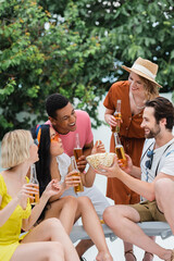 smiling man holding bowl of popcorn near cheerful interracial friends drinking beer