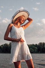 Fototapeta premium A young woman in a hat smiles. A beautiful happy blonde in a white summer dress and a straw hat on her head is standing in the water and posing against the background of trees. 