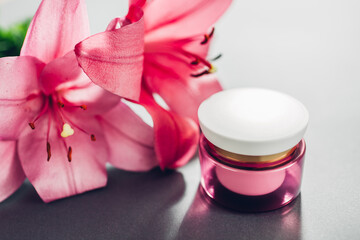 Obraz na płótnie Canvas Jar with moisturizer cream surrounded with pink lily flowers on grey background. Organic cosmetics