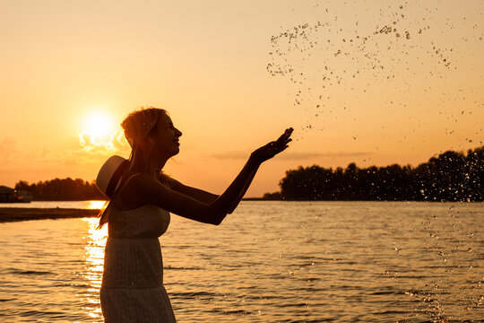 Photo Of A Young Woman At Sunset. Situ Is A Side View Of A Slender, Beautiful Blonde In A Summer Dress And A Straw Hat Standing In The Water And Splashing Water. 