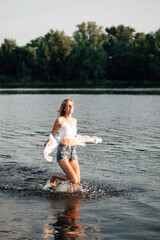A beautiful woman runs freedom through the water. A smiling young blonde in a white top and shirt and denim shorts against a landscape background. 